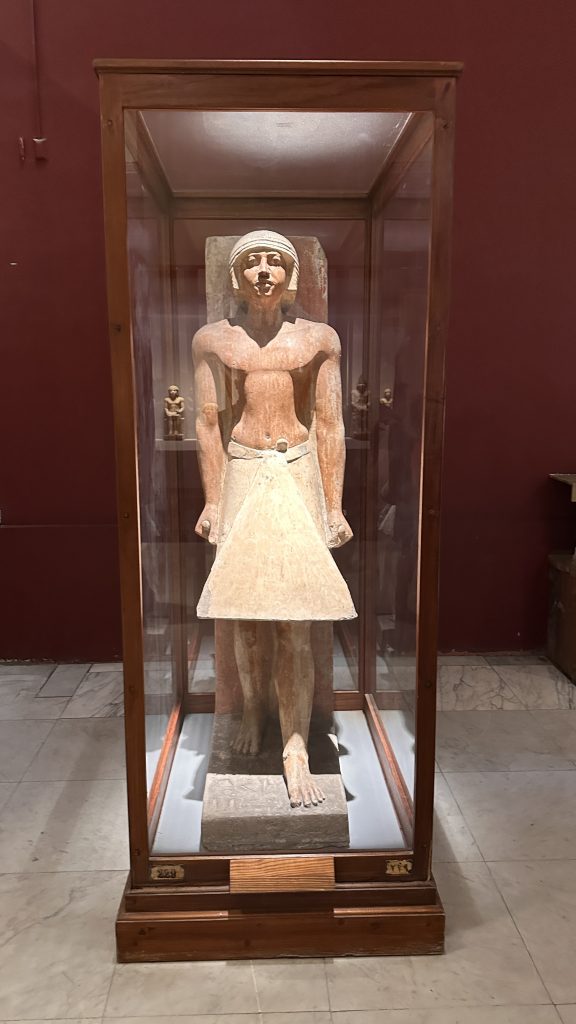 Painted ancient Egyptian statue of a man standing in a wooden-framed glass display case at the Egyptian Museum in Cairo on Day 3 of a 10-day Egypt itinerary.
